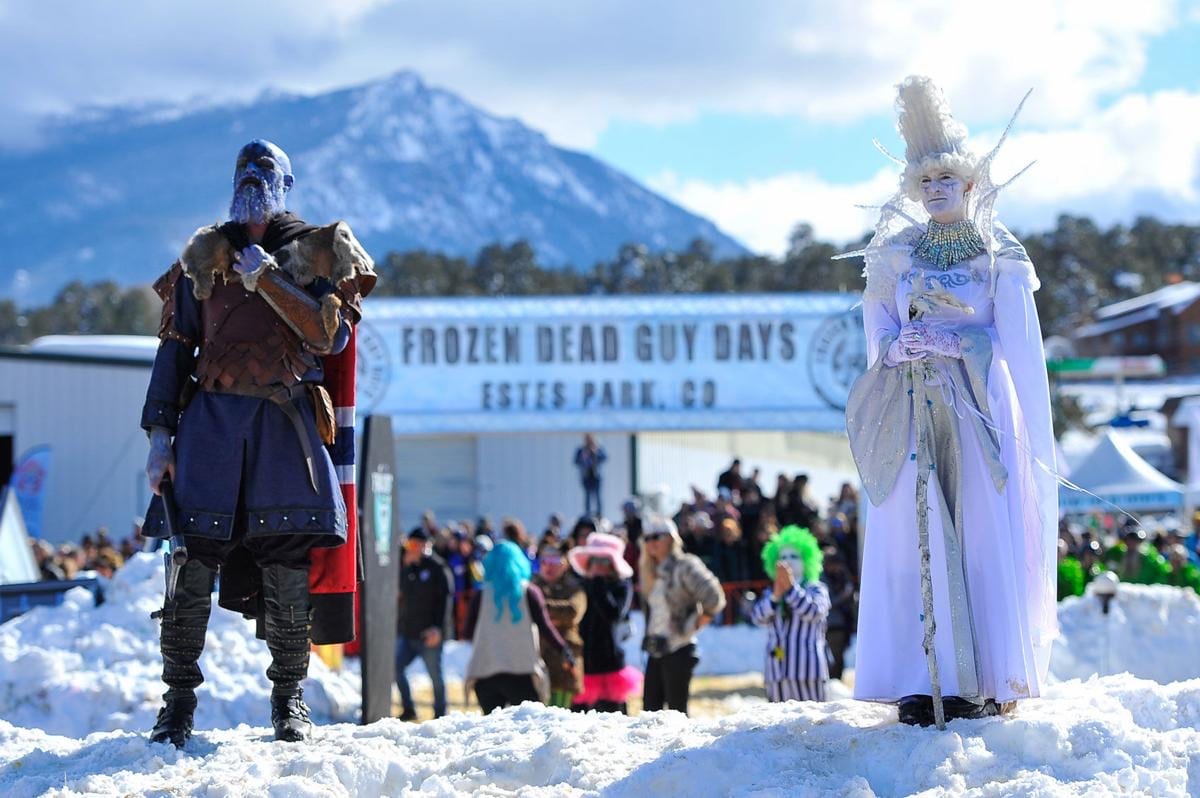 A Viking in blue face paint and an Ice Queen in white stand on snowbanks before a Frozen Dead Guy Days banner, a snowy peak behind.