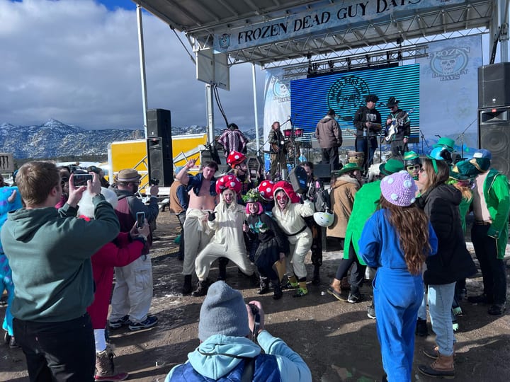 A costumed crew in mushroom hats, onesies and leprechaun green poses in front of the outdoor stage while a band plays.
