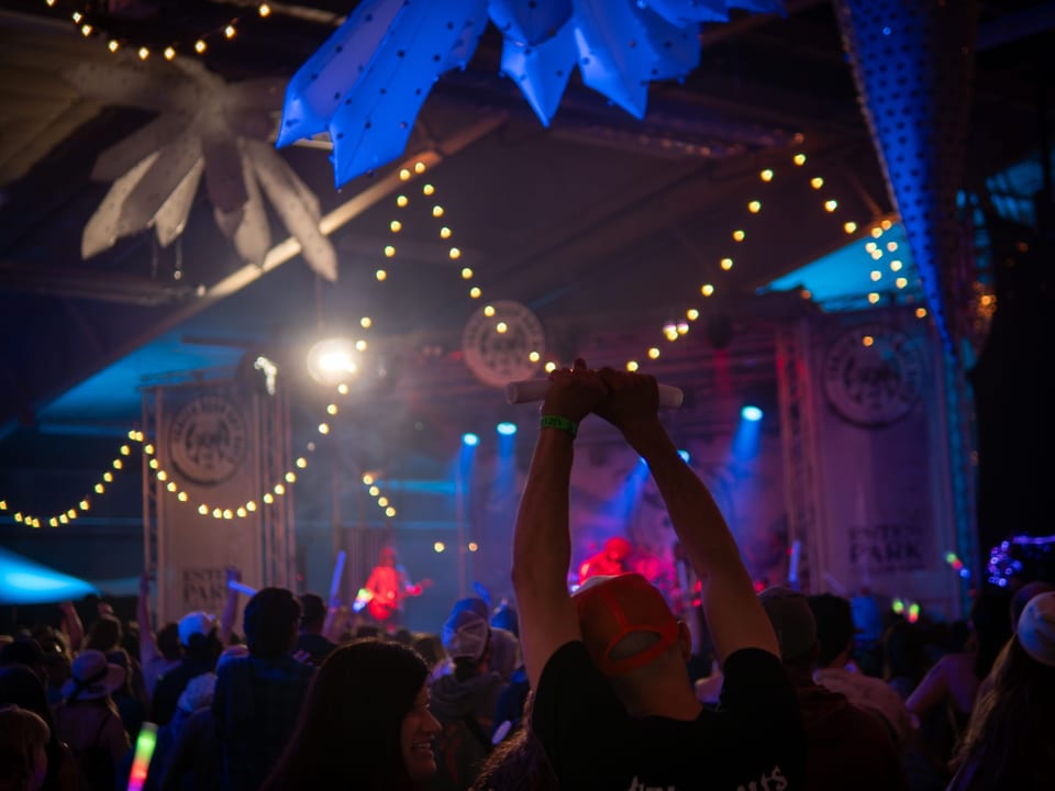 A fan raises both arms with a glow stick under string lights, a band on a red-lit stage beyond.