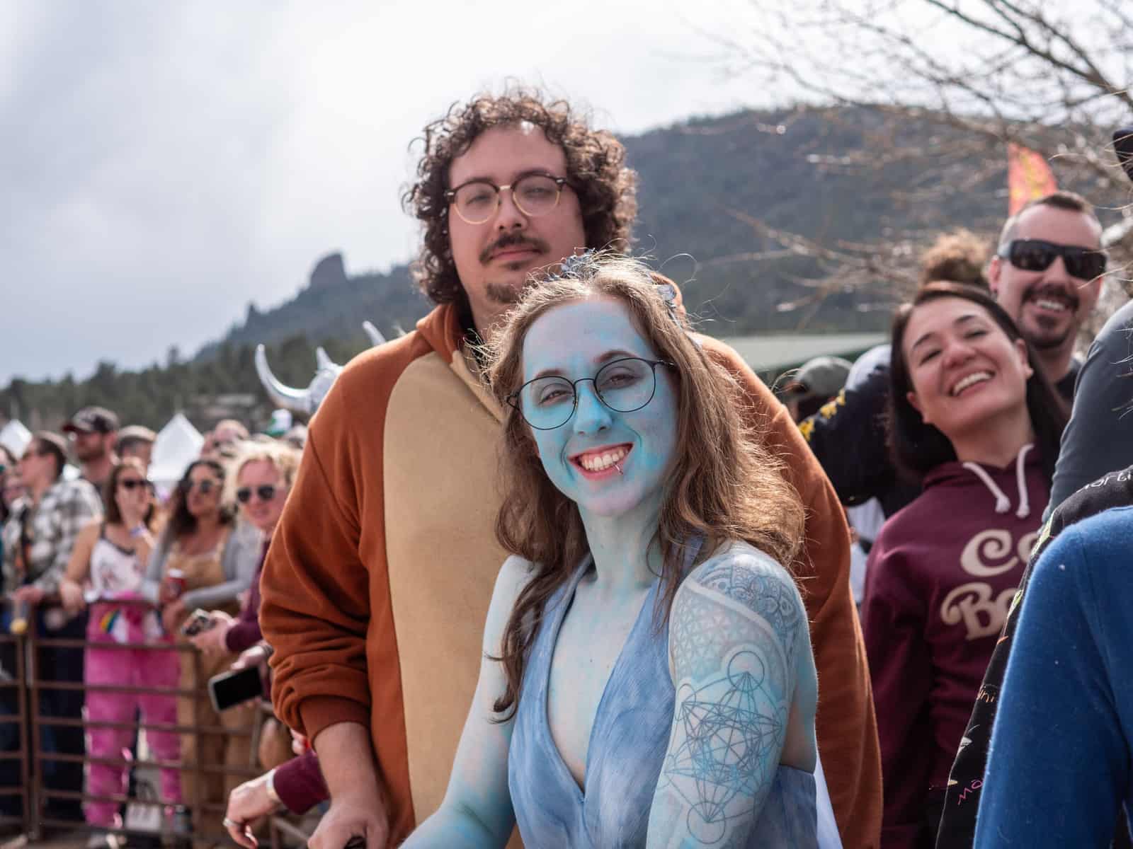 A smiling woman in blue body paint and glasses stands with a friend in a bear costume at the race rail.