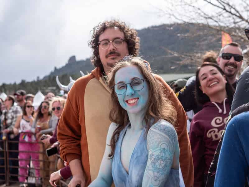 A smiling woman in blue body paint and glasses stands with a friend in a bear costume at the race rail.