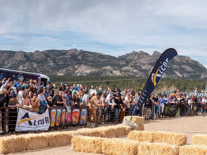 Spectators pack the coffin-course rail beside an Alcor flag and hay bales, rocky peaks and wide sky behind.