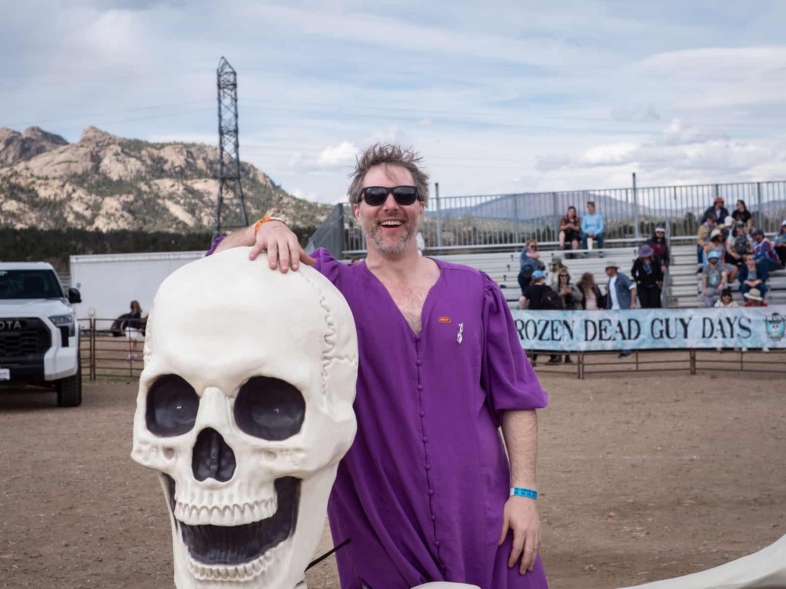 A grinning man in a purple robe rests his hand on a giant skull prop in front of a Frozen Dead Guy Days banner.