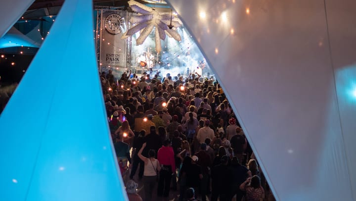 Seen past a glowing ice wall, a crowd fills the hall in front of a smoky, lit stage.