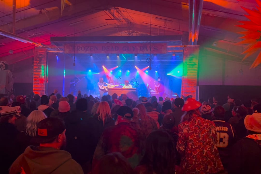 Coloured beams sweep over a costumed crowd facing the Frozen Dead Guy Days stage banner.