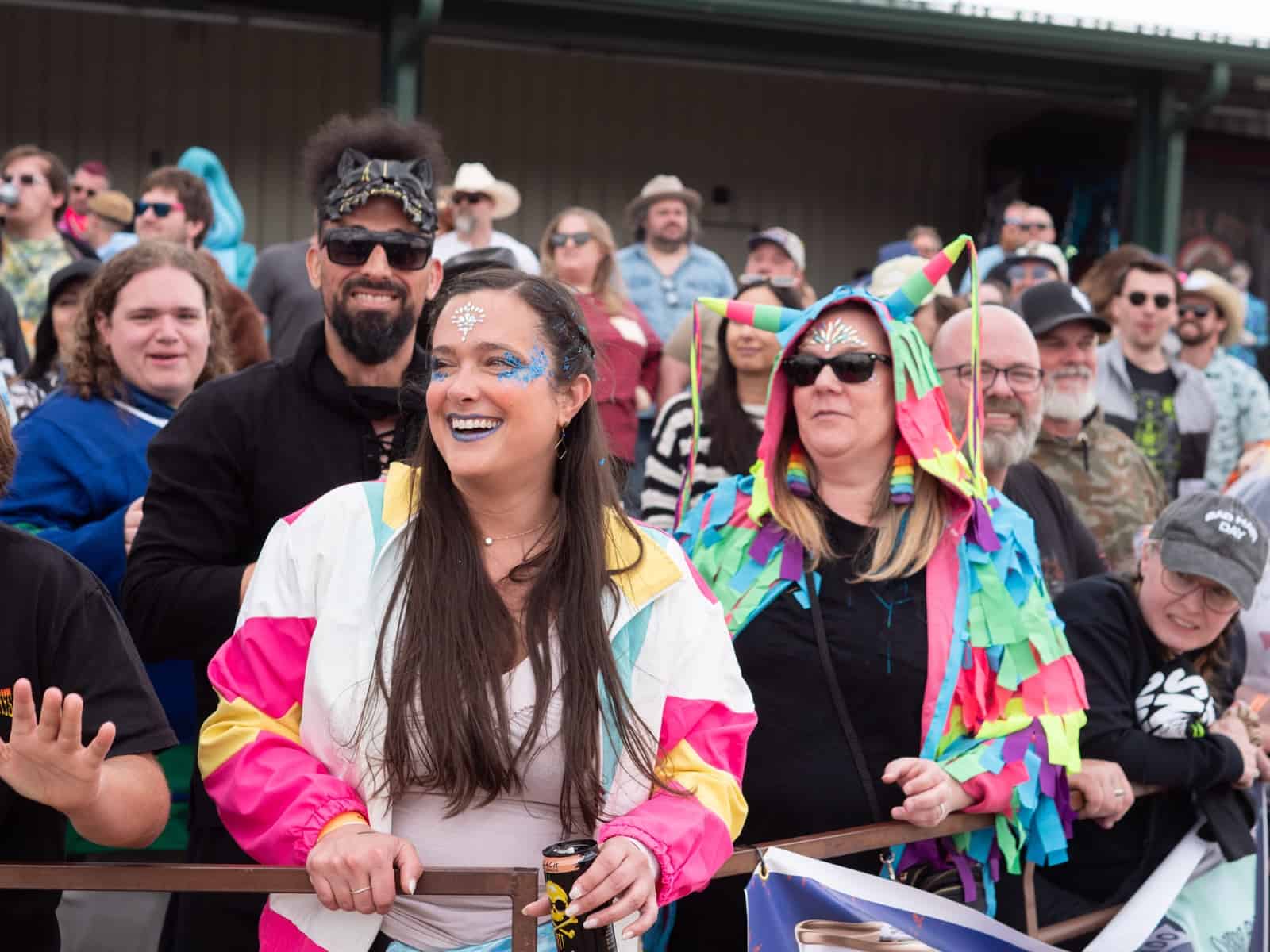 Fans laugh and wave at the race rail, a woman in a bright windbreaker and face gems up front.