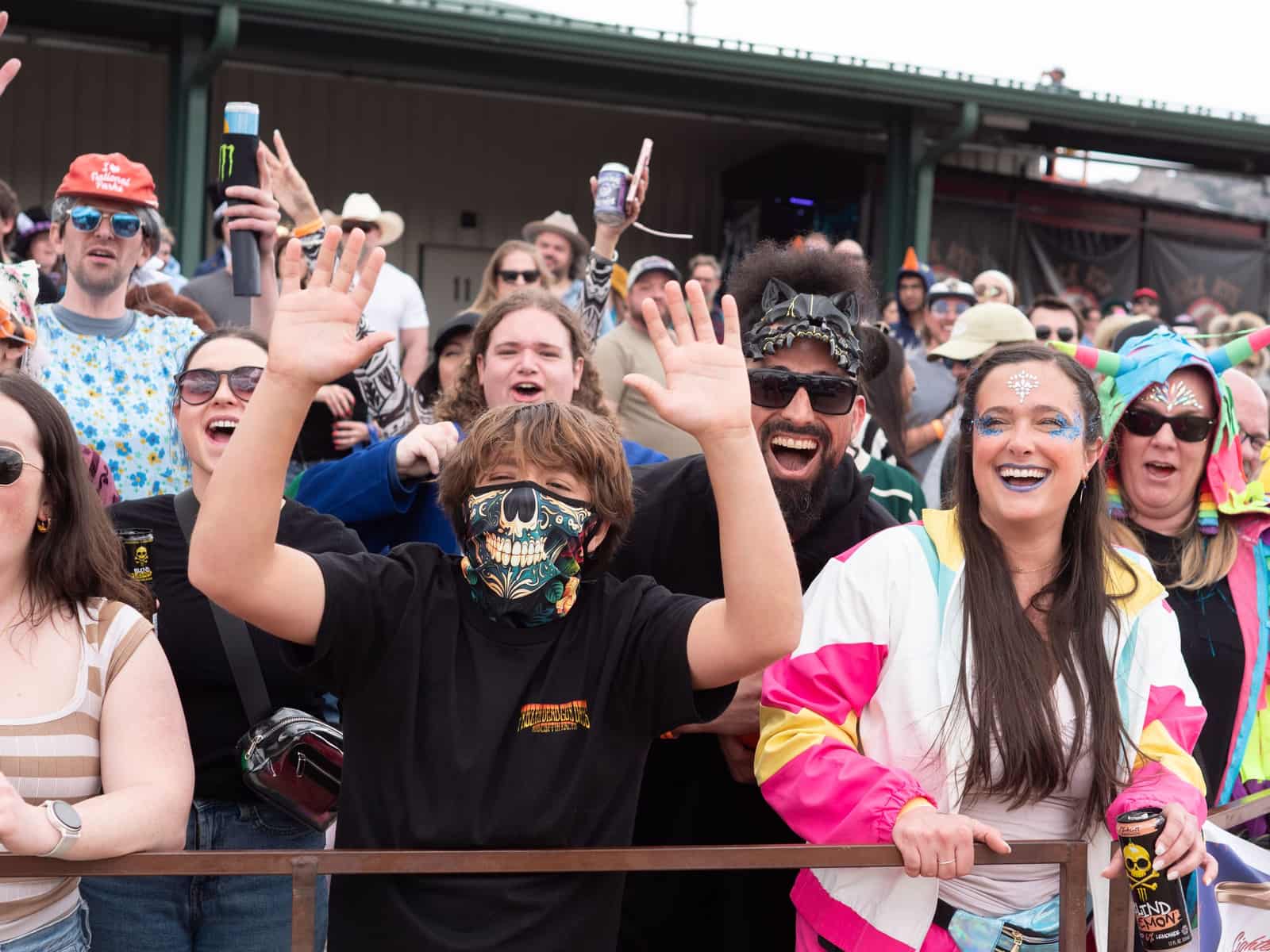 Fans cheer and wave at the race rail: a young fan in a skull-print neck gaiter up front, a woman in a bright 80s windbreaker with face gems beside him.