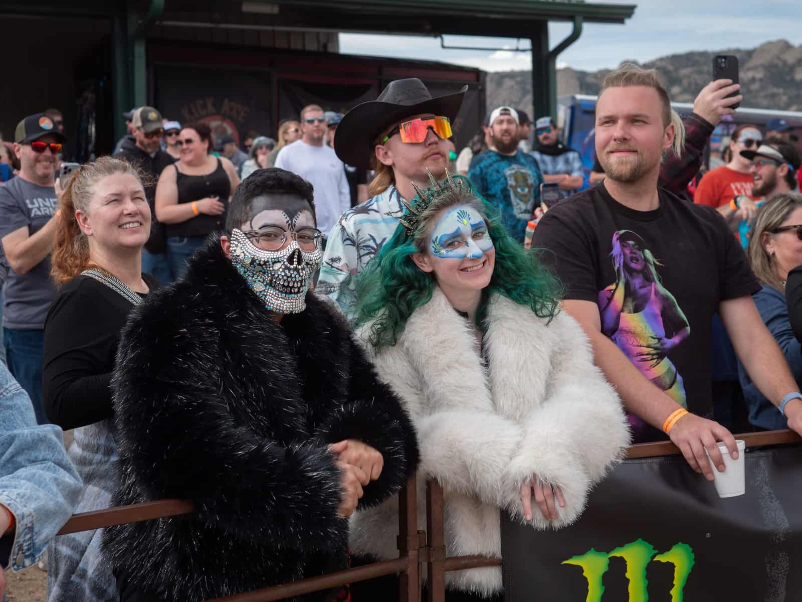 A fan in a sequined skull mask and a green-haired fan with ice-blue face paint lean on the race rail.