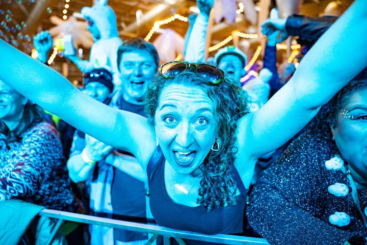 A woman at the front rail throws her arms up and shouts with joy, surrounded by cheering fans under string lights.