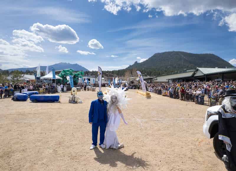 Two festival characters, one in blue face paint, a crown and a blue suit, the other an Ice Queen in white, stand in the race arena under a huge blue sky.