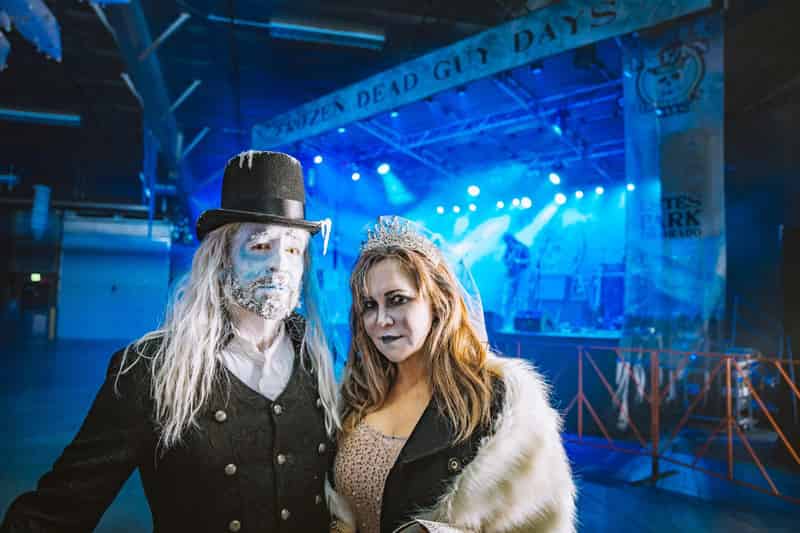 A Grandpa Bredo look-alike with a frosted beard and top hat and a frozen bride in a white fur stole pose on the festival stage under blue lights.