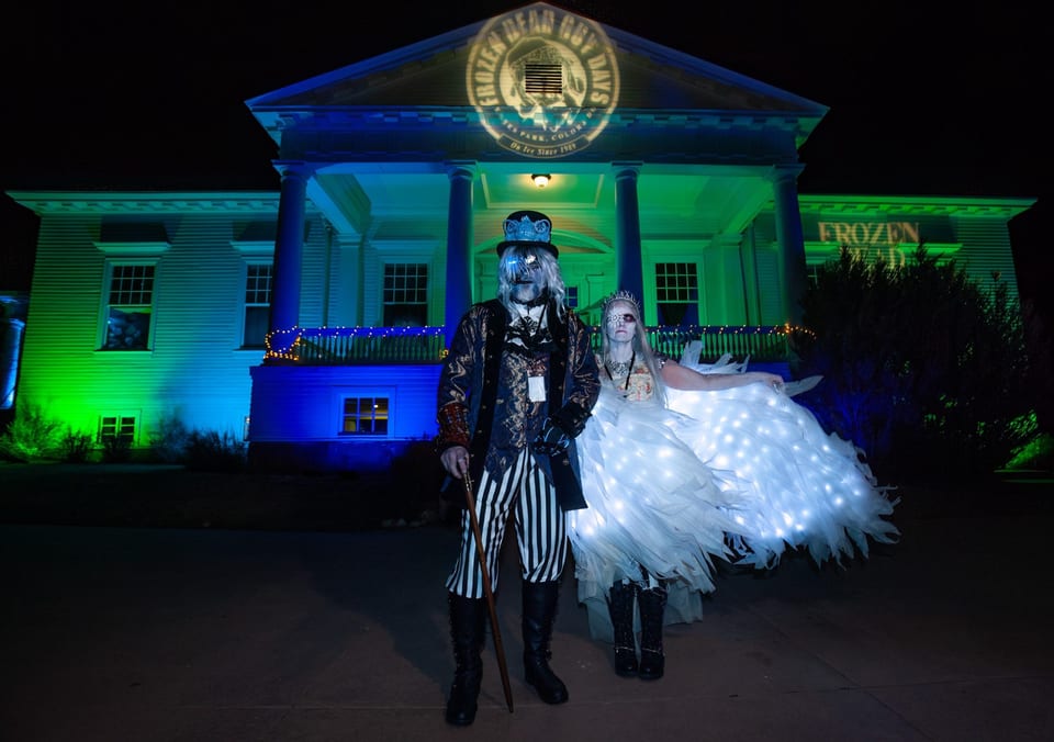 A frozen grandpa in a skull top hat and a bride with glowing wings pose outside the ballroom at night, the festival badge projected above.
