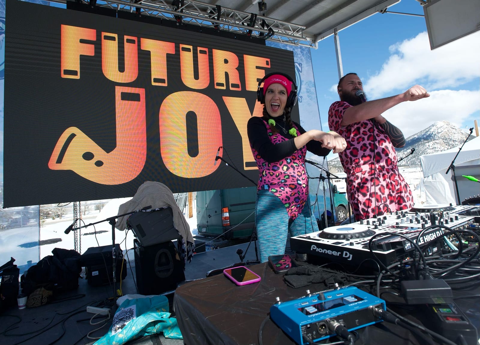 DJ duo Future Joy dance in leopard jumpsuits on the outdoor stage, a snowy peak behind.