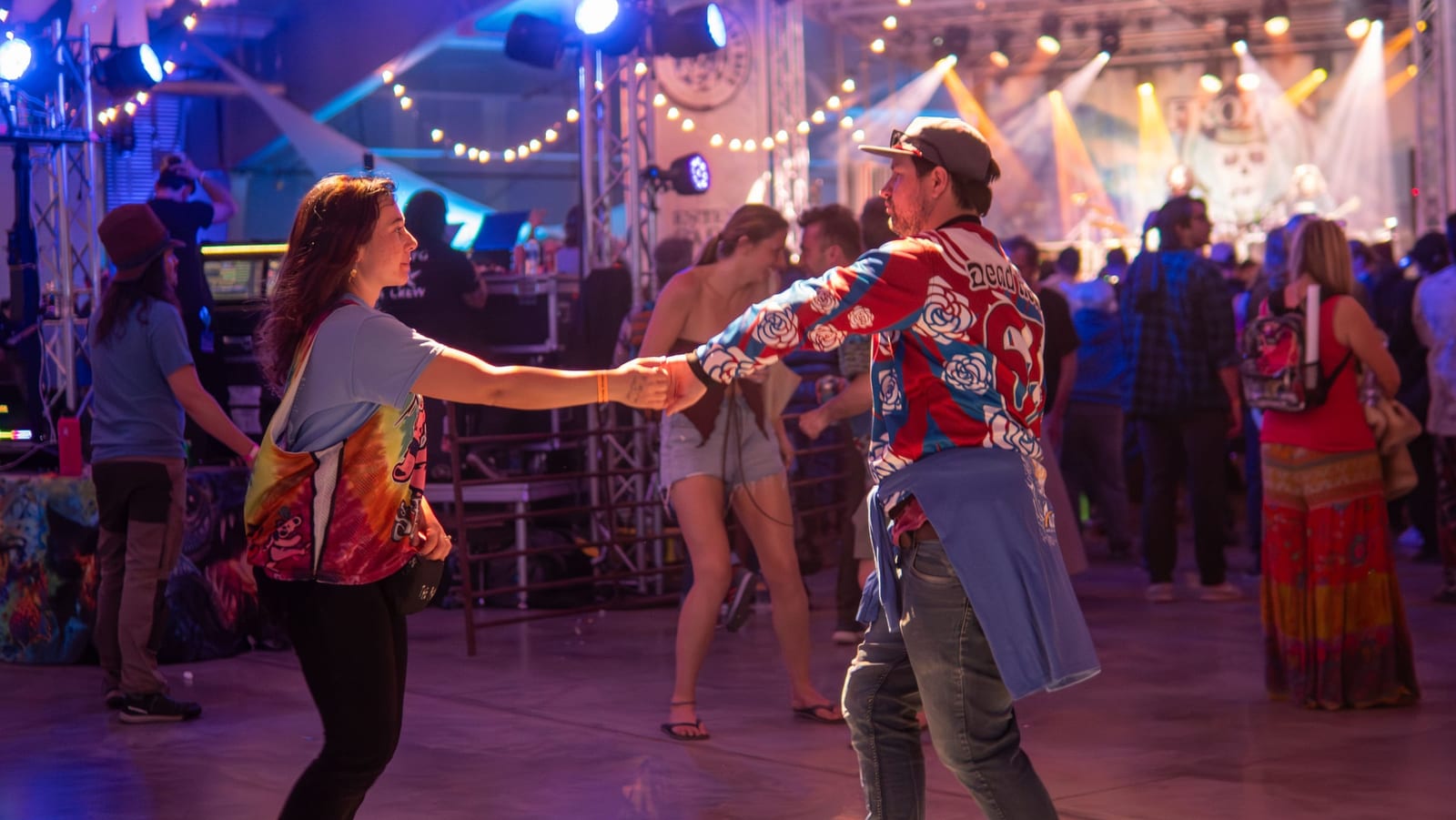 A couple swing-dances hand in hand in front of a glowing stage with string lights, one in a rose-print jacket.