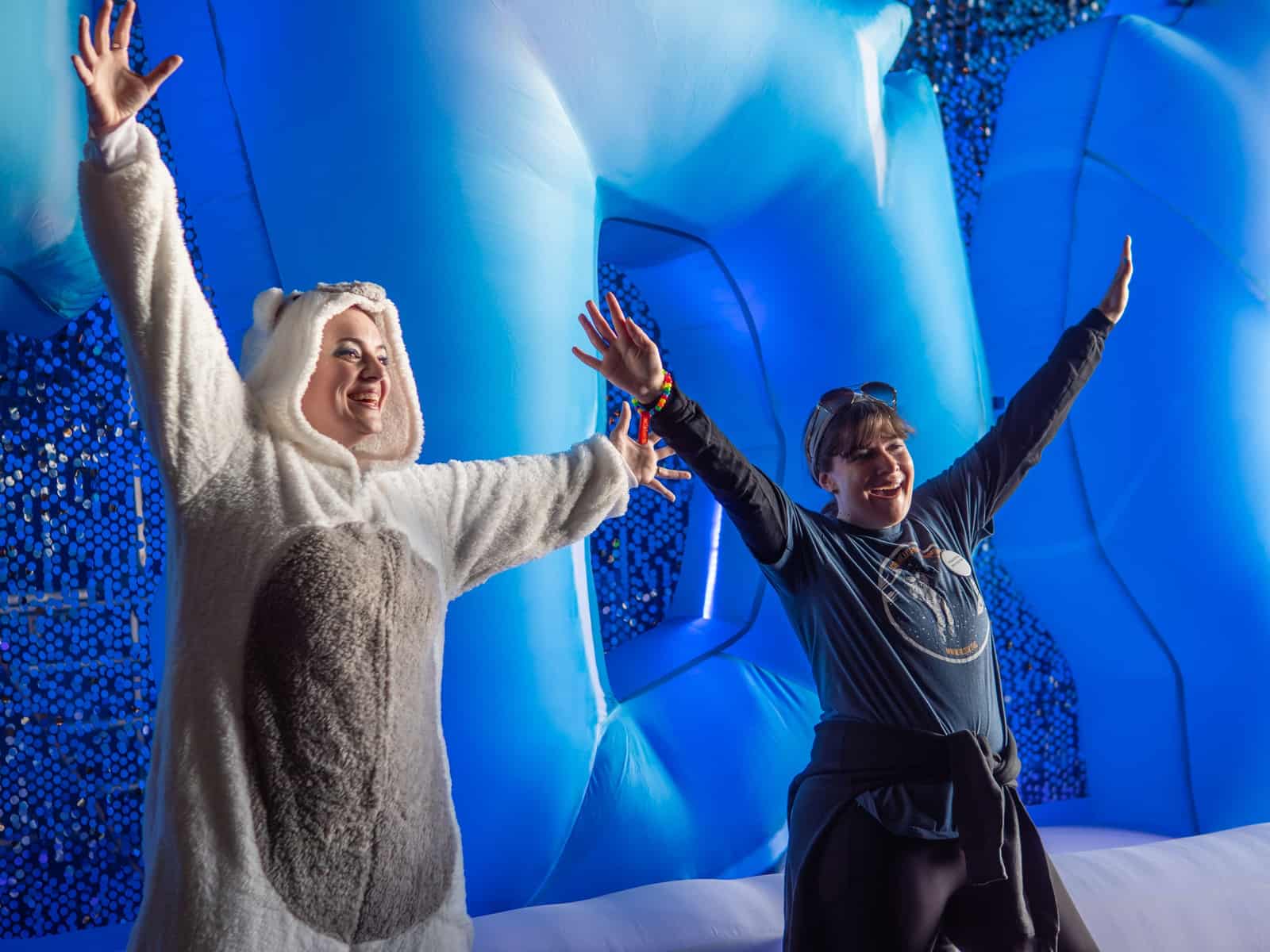 Two women throw their arms wide inside a glowing blue ice-cave inflatable, one in a polar bear onesie.