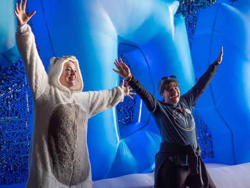 Two women throw their arms wide inside a glowing blue ice-cave inflatable, one in a polar bear onesie.