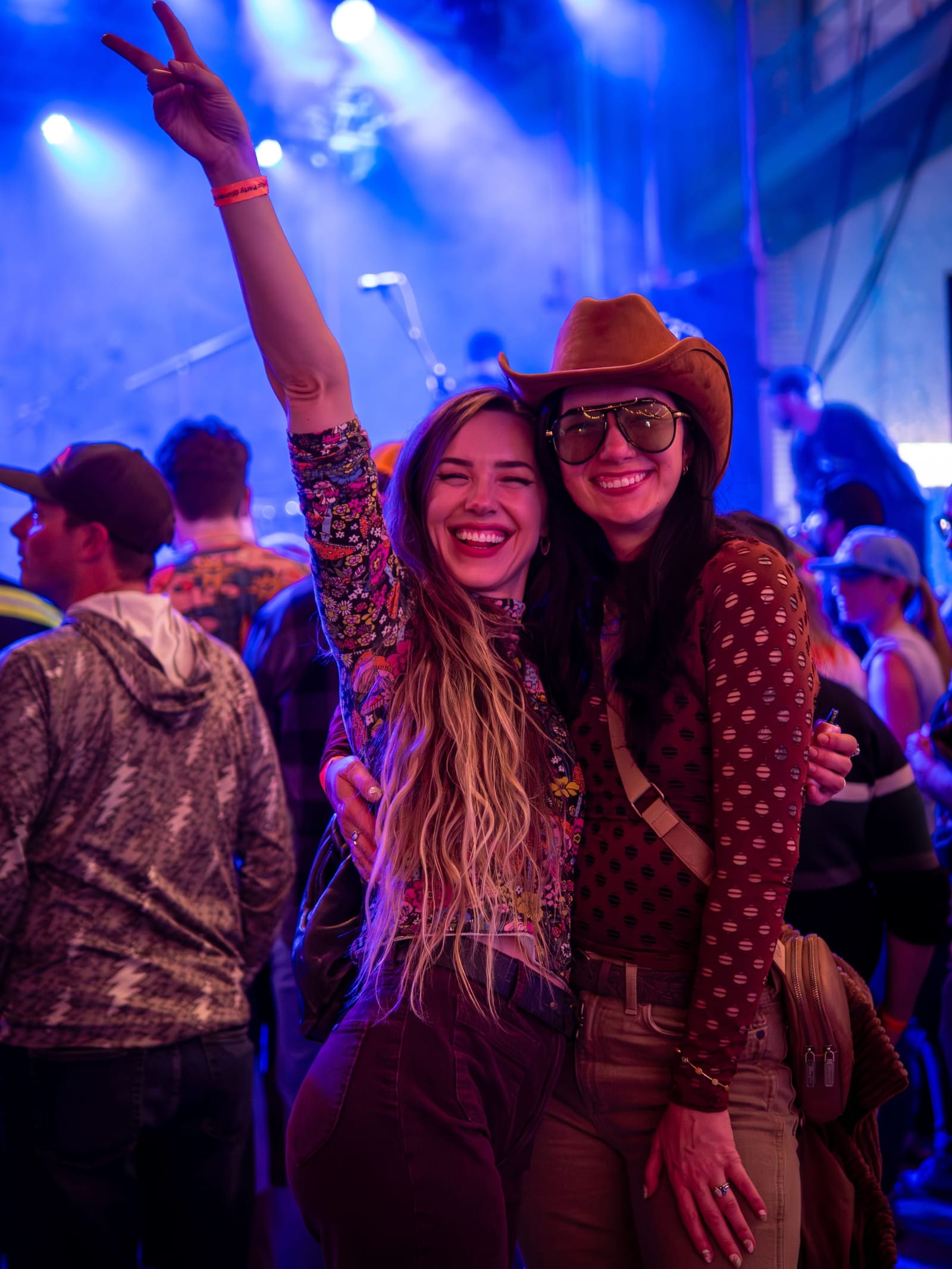 Two smiling friends, one throwing a peace sign and one in a cowboy hat, under blue concert lights.