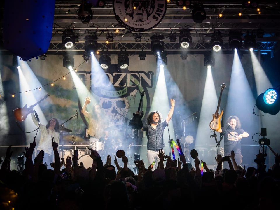 Pigeons Playing Ping Pong raise their arms under white spotlights as the crowd's raised hands fill the foreground.