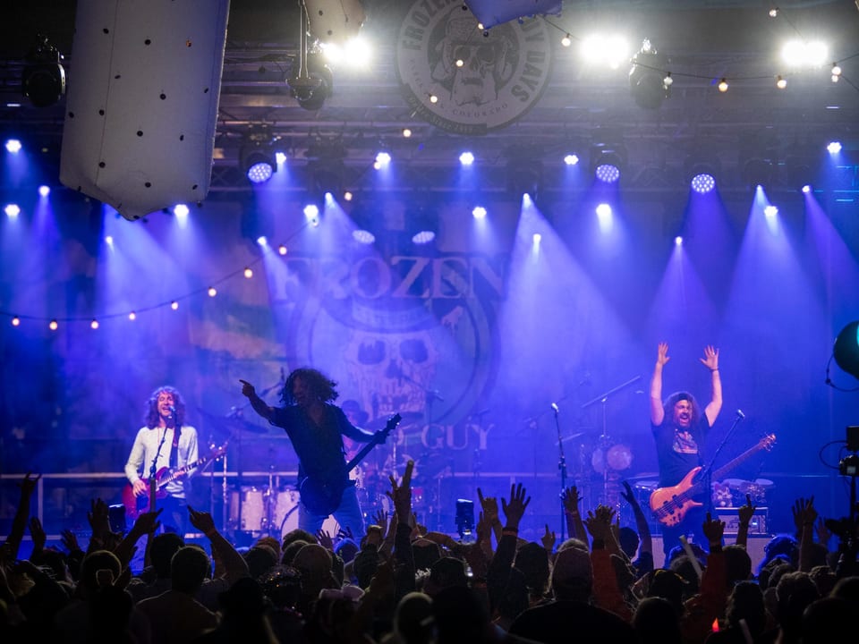 Pigeons Playing Ping Pong play under blue lights, one guitarist pointing to the crowd and the bassist with both arms raised.