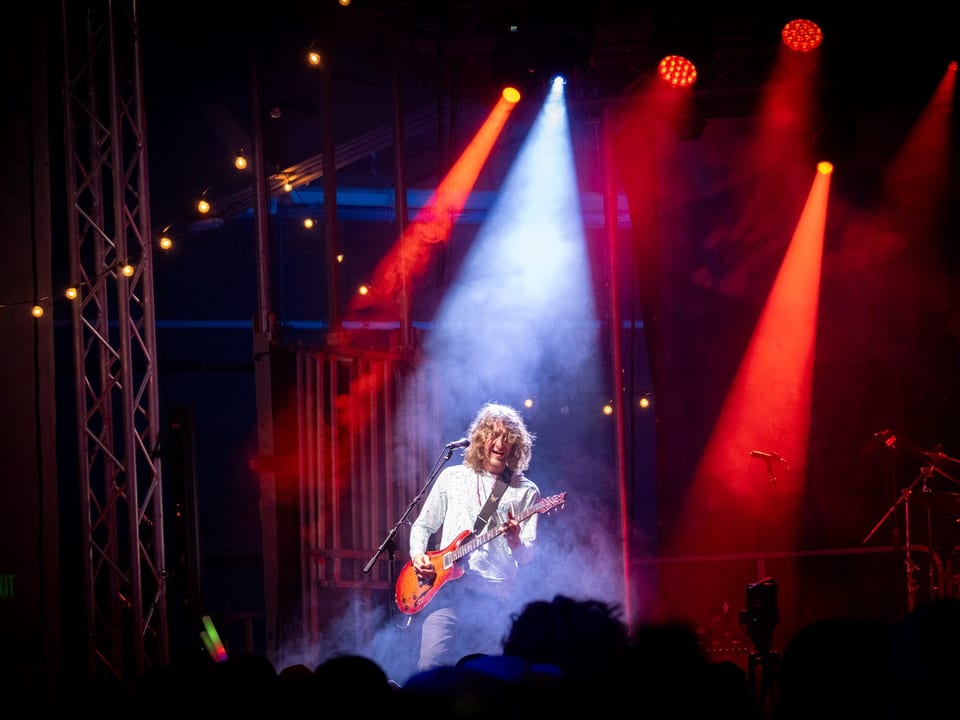 A guitarist solos in a white beam between red spotlights, haze drifting across the dark stage.