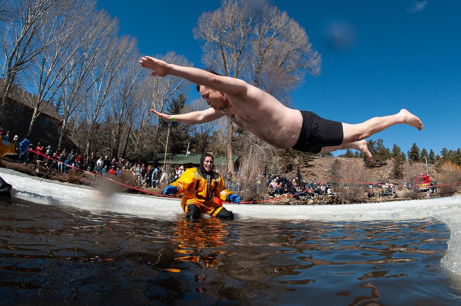 A plunger dives flat out over an icy pool as a rescue swimmer waits in the water, a crowd and pine trees behind.