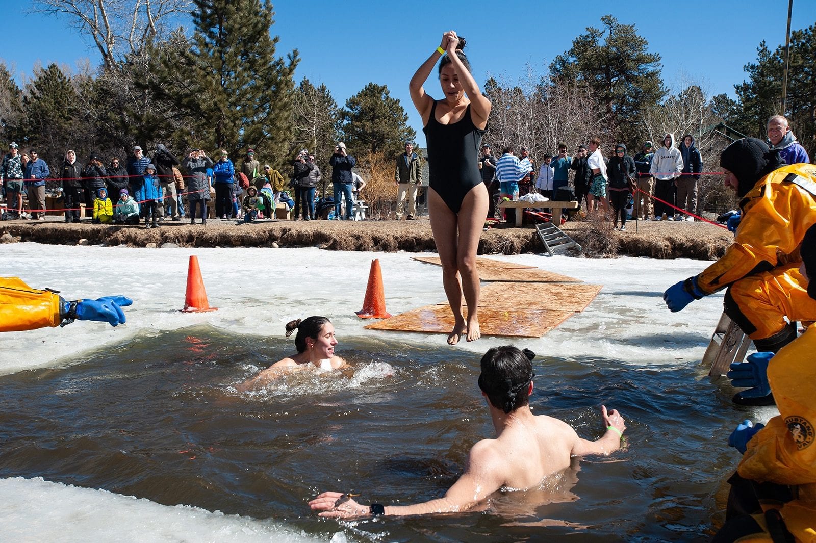 A plunger in a swimsuit raises her arms at the edge of an icy pool while two others swim below, a crowd watching from the snowbank.