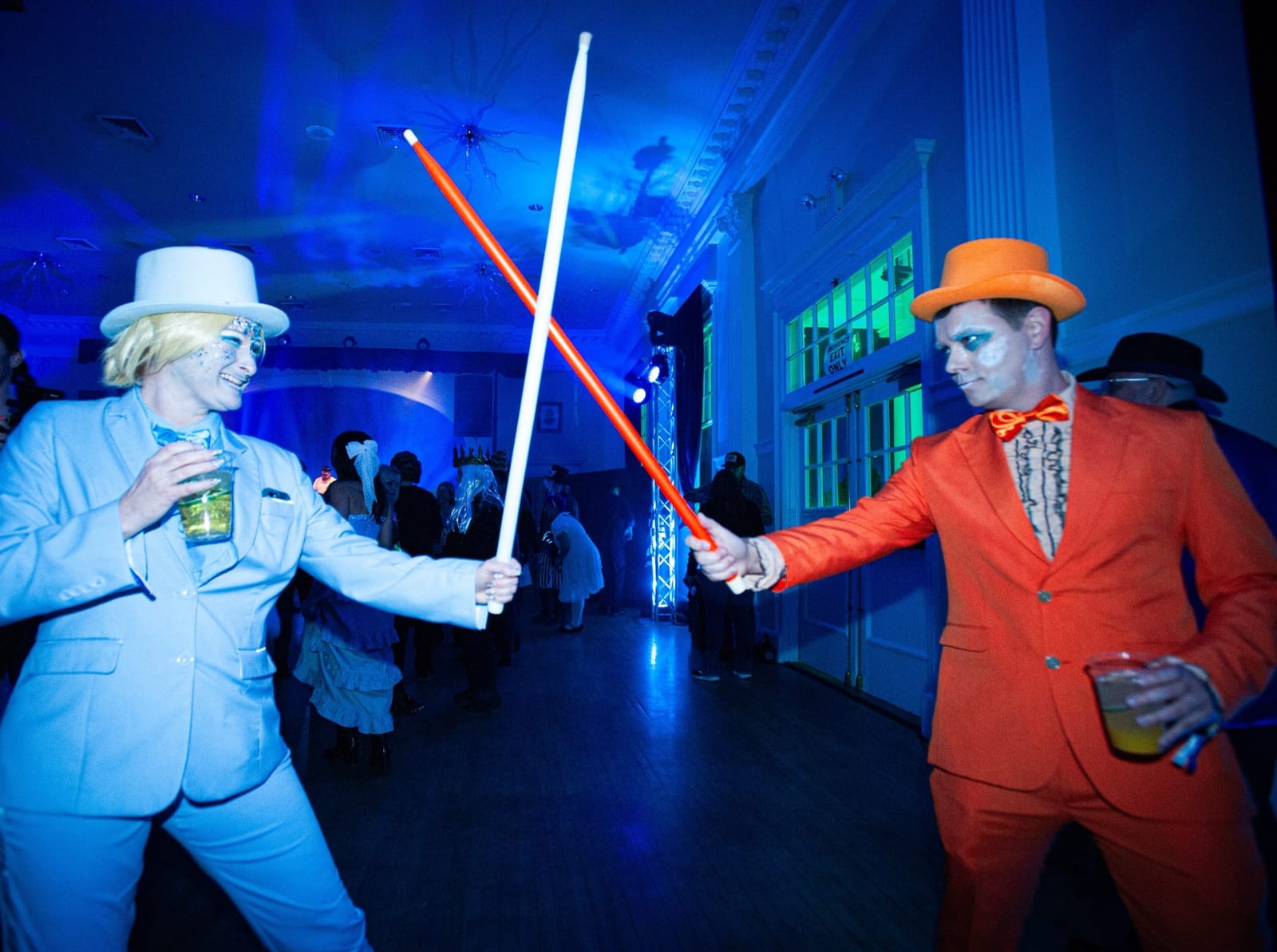 Two guests in powder-blue and orange tuxedos duel with light sabres on the ballroom floor.