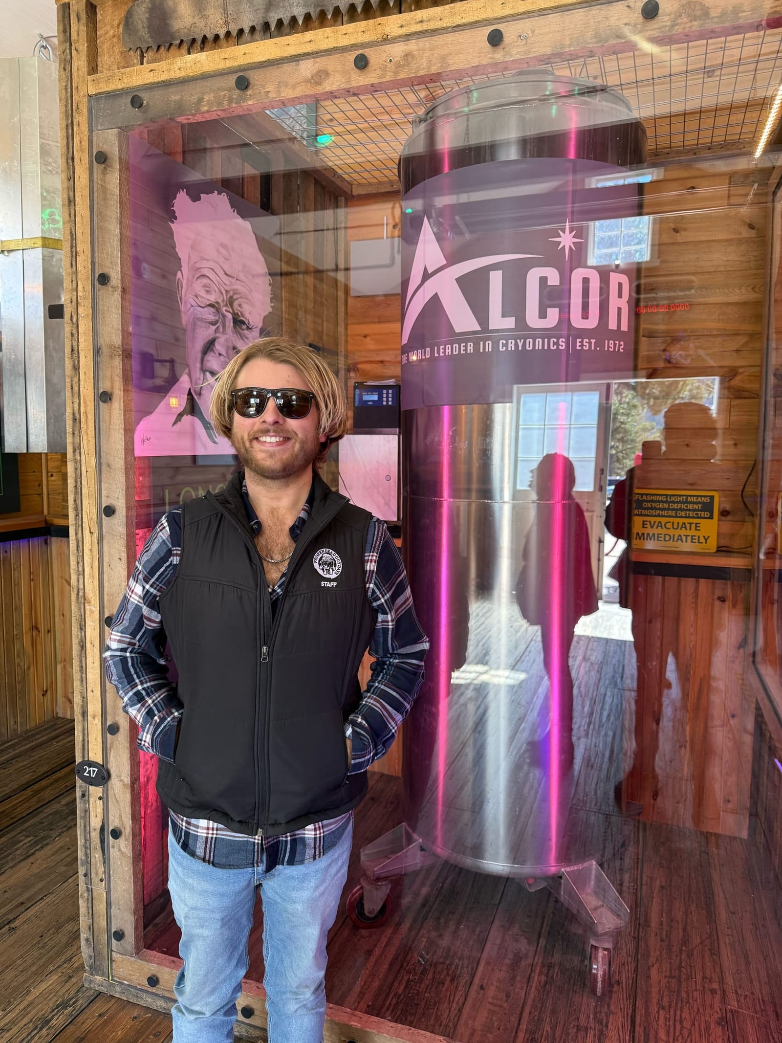 A smiling festival staffer in sunglasses stands before a cryonics capsule in Alcor's museum exhibit.