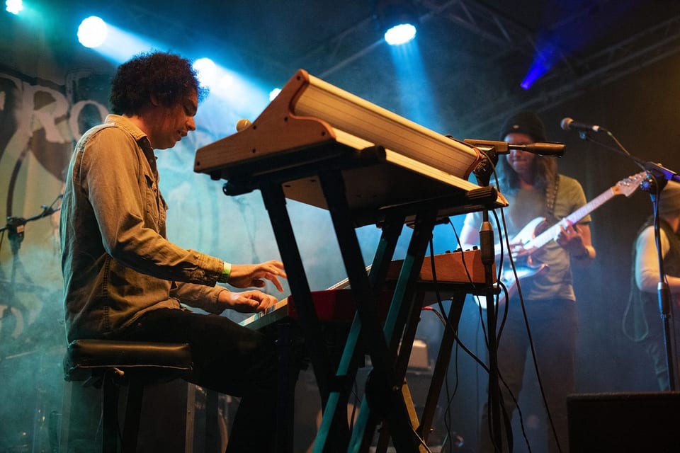 A curly-haired keyboardist plays in teal light, a guitarist behind him.