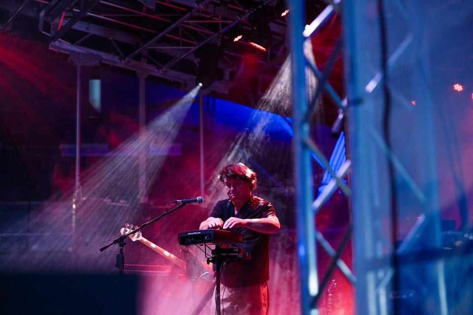 A musician plays keys and sings beside the stage truss in red and purple light.