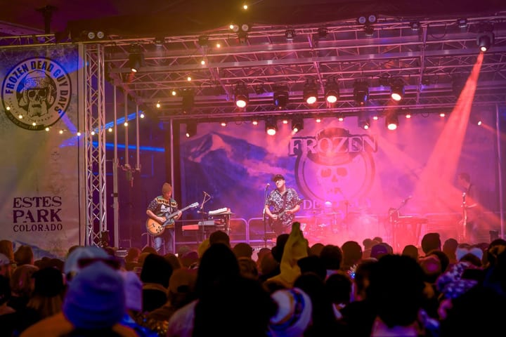 Two guitarists play in pink haze before a Frozen skull backdrop and an Estes Park banner.
