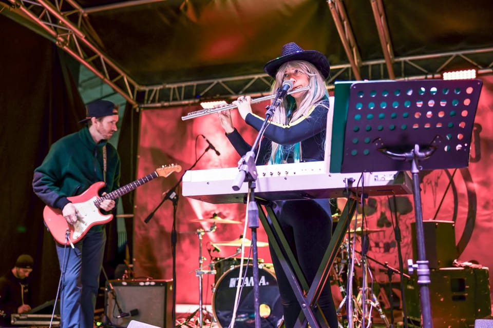 A musician in a cowboy hat plays flute at a keyboard beside a guitarist on a red-lit stage, the drummer behind them.