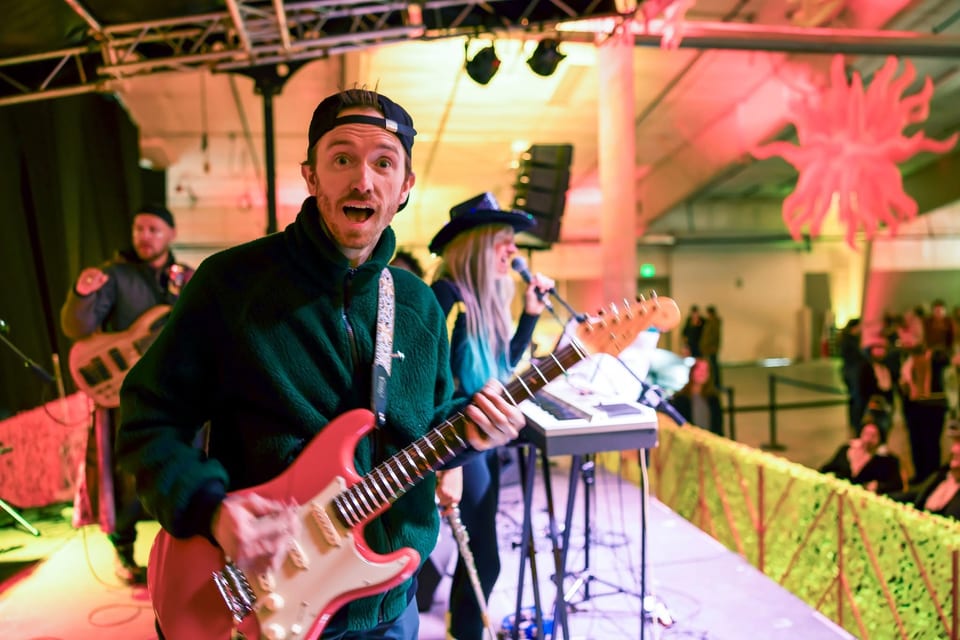 A guitarist in a backwards cap grins wide at the camera mid-song, a singer in a cowboy hat at the keyboard behind him.
