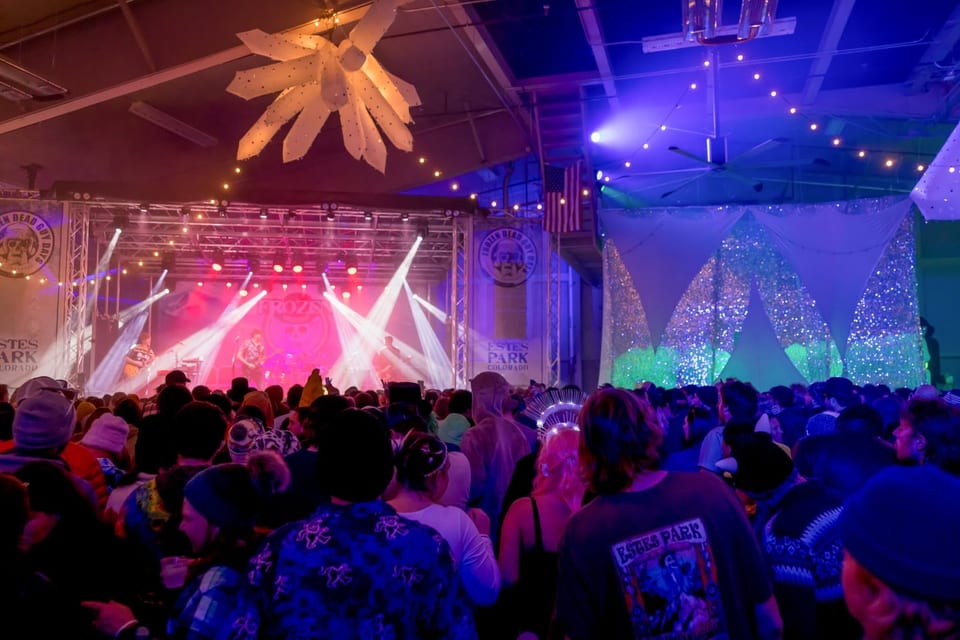 A band plays under pink and white light beams as a packed crowd watches, ice crystals hanging overhead.
