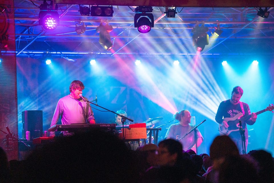A keyboard player sings and a bassist plays under sweeping blue and pink stage beams, fans' heads silhouetted in front.