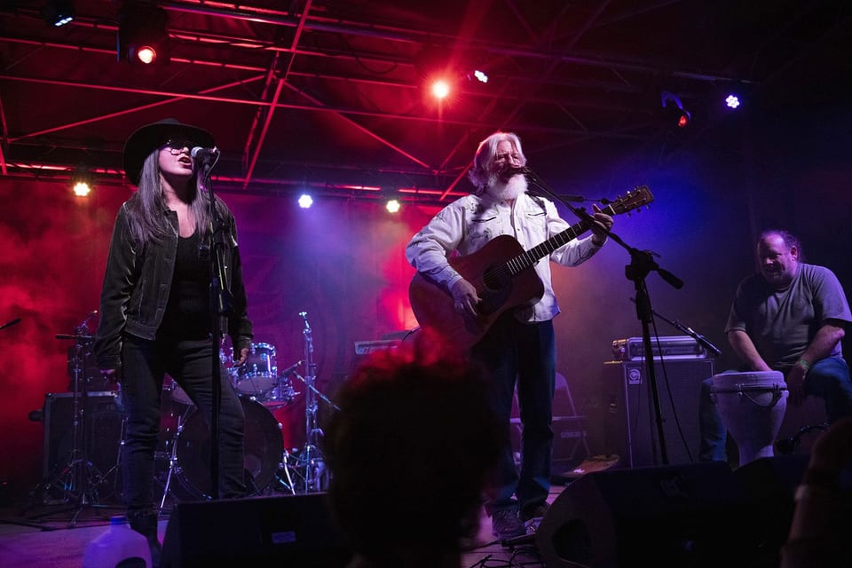 A singer in a black cowboy hat and a white-bearded acoustic guitarist perform under red and purple light, a hand drummer beside them.