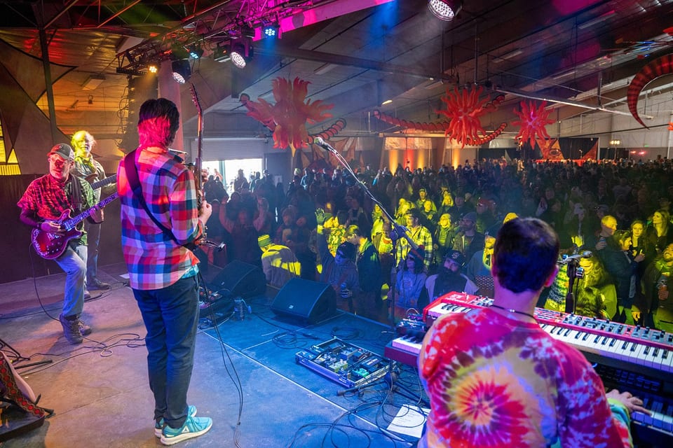 Seen from the stage behind two guitarists and a keyboard player, a packed hall of fans dances under yellow and red light.