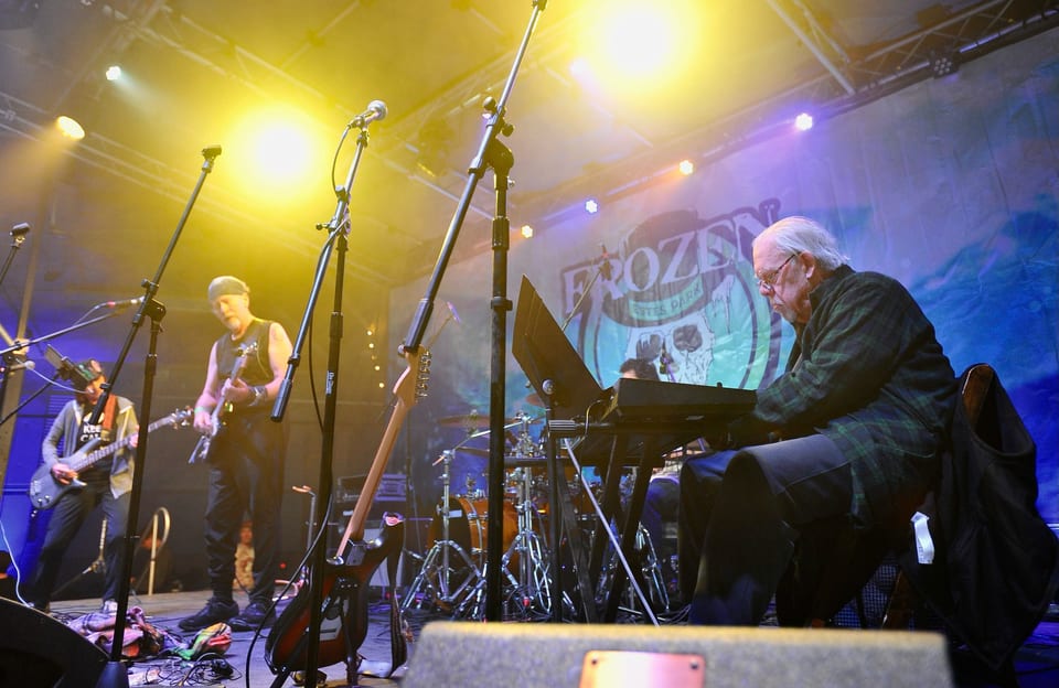 A white-haired keyboardist and two guitarists play under bright yellow stage light in front of the Frozen backdrop.