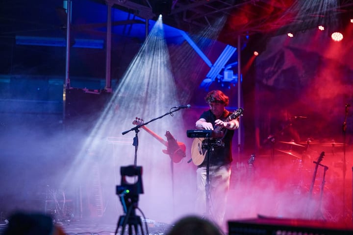 A curly-haired musician plays guitar and a small keyboard as a white spotlight beam cuts through pink and blue stage fog.