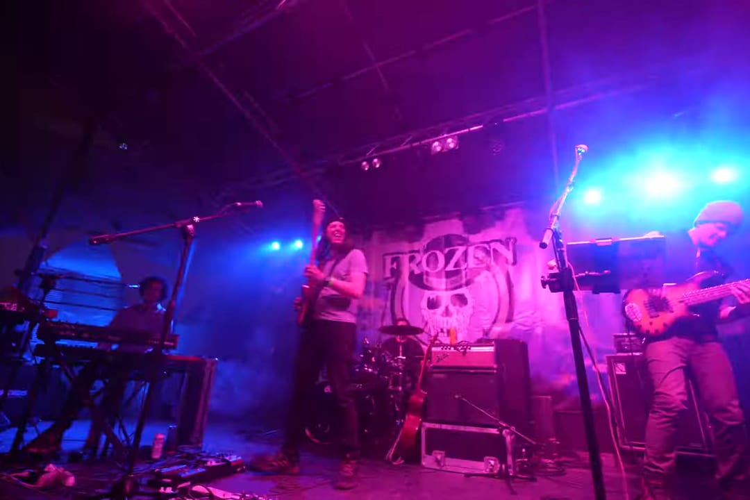 A band plays in pink and blue light before a Frozen Dead Guy Days skull backdrop.