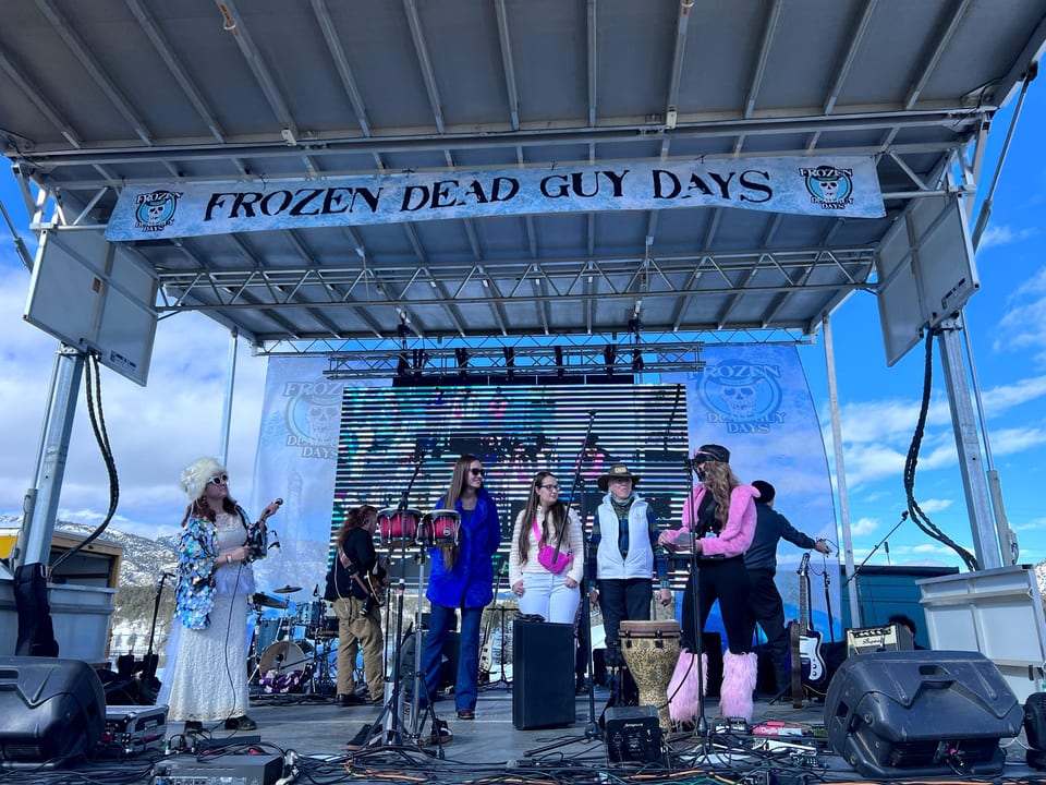 A costumed band in a white fur hat, sequins and pink fuzzy boots performs on the outdoor Frozen Dead Guy Days stage under blue sky.