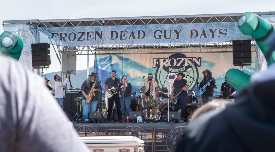 A big band with a saxophone plays the outdoor stage under the Frozen Dead Guy Days banner, a coffin on hay bales in front.