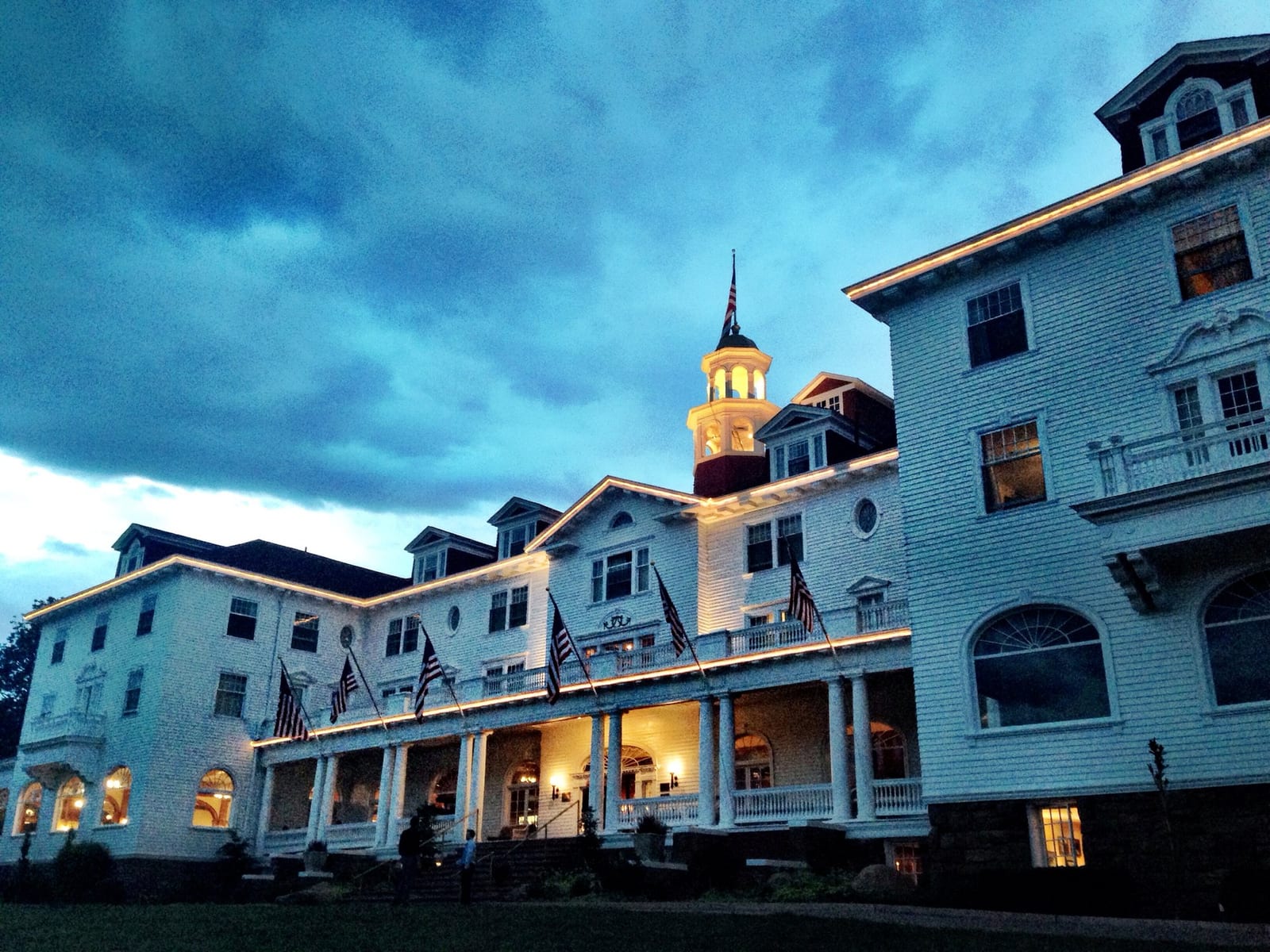 The Stanley Hotel lit up at dusk: warm light along the roofline and columned porch, the white cupola glowing against a dark, stormy blue sky.