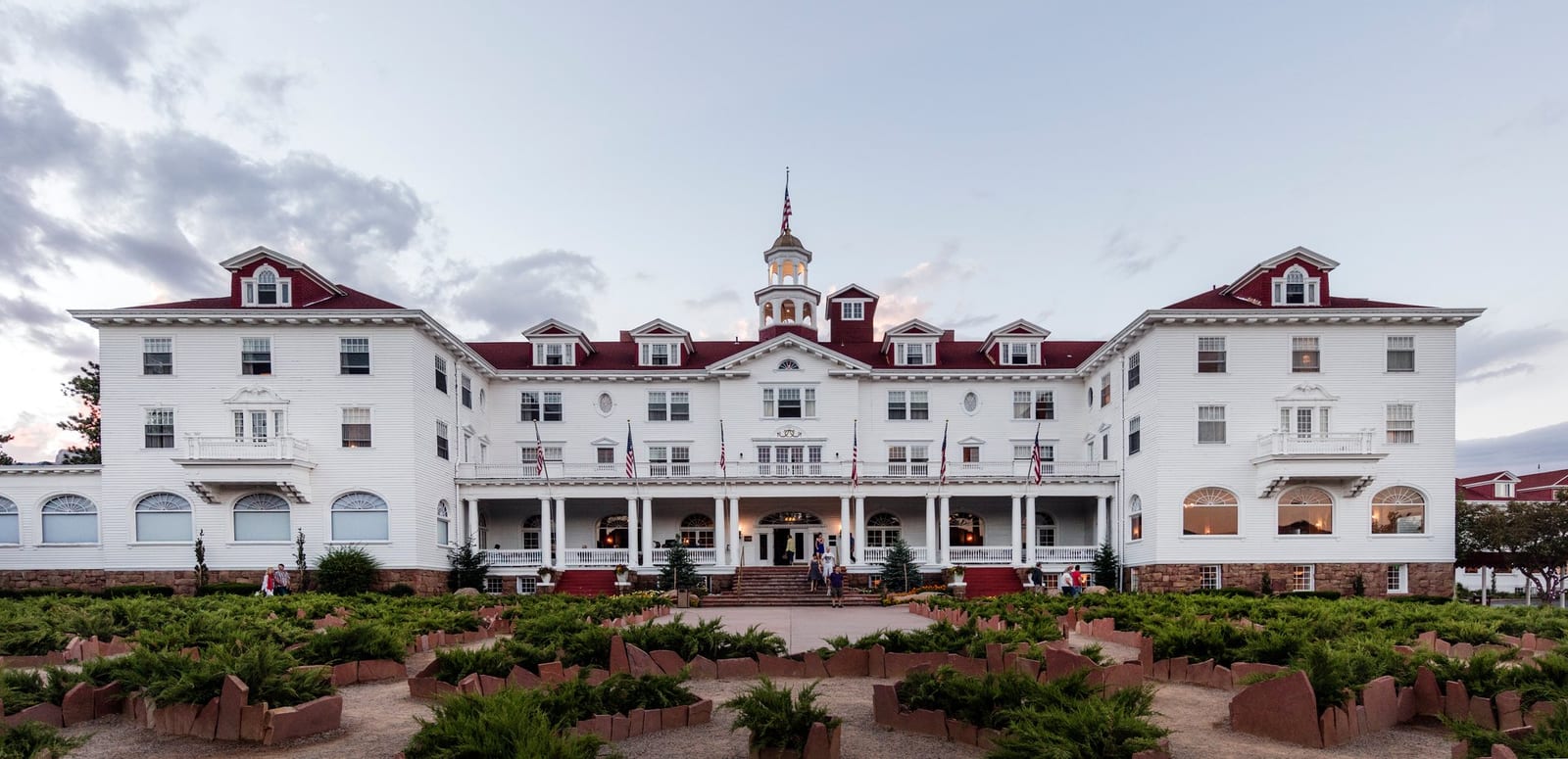 The Stanley Hotel at dusk: a long white facade with red roofs, a central cupola and a columned front porch, seen across a terraced garden.