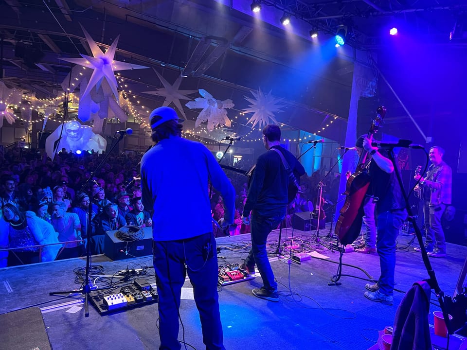 From behind Yonder Mountain String Band on stage toward a packed rail under star inflatables and string lights.